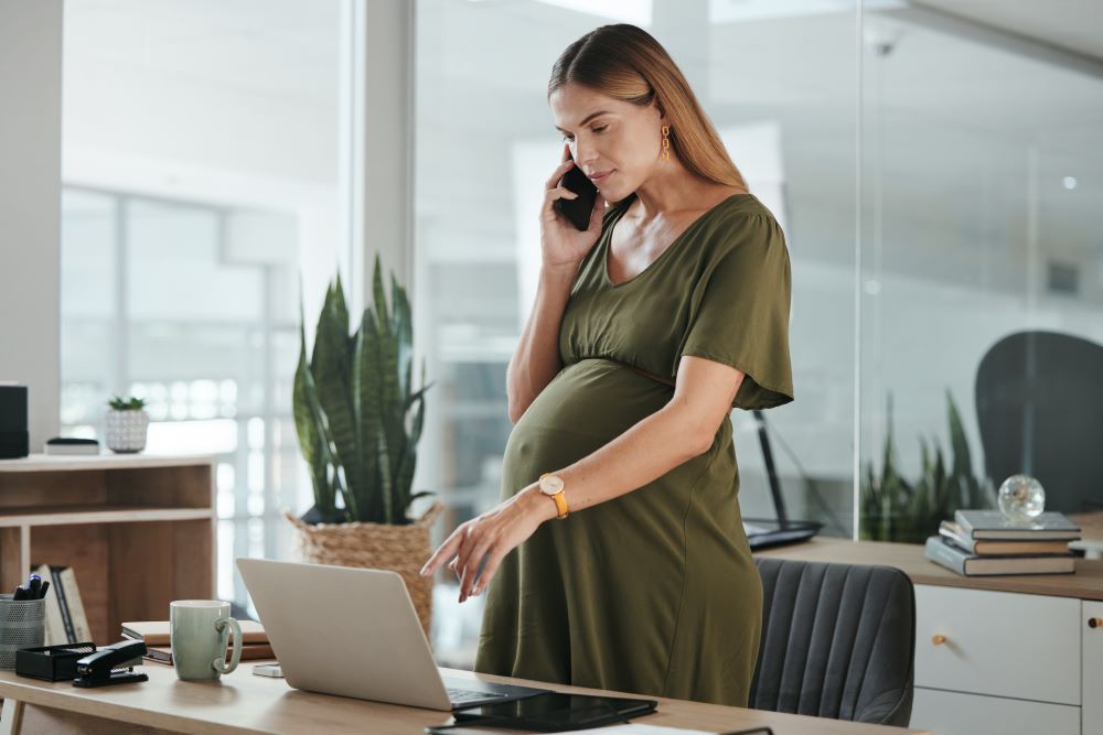 Gestante em ambiente corporativo, de pé ao lado de notebook, ao telefone — representando a realidade do RH diante da estabilidade gestante prevista na CLT.