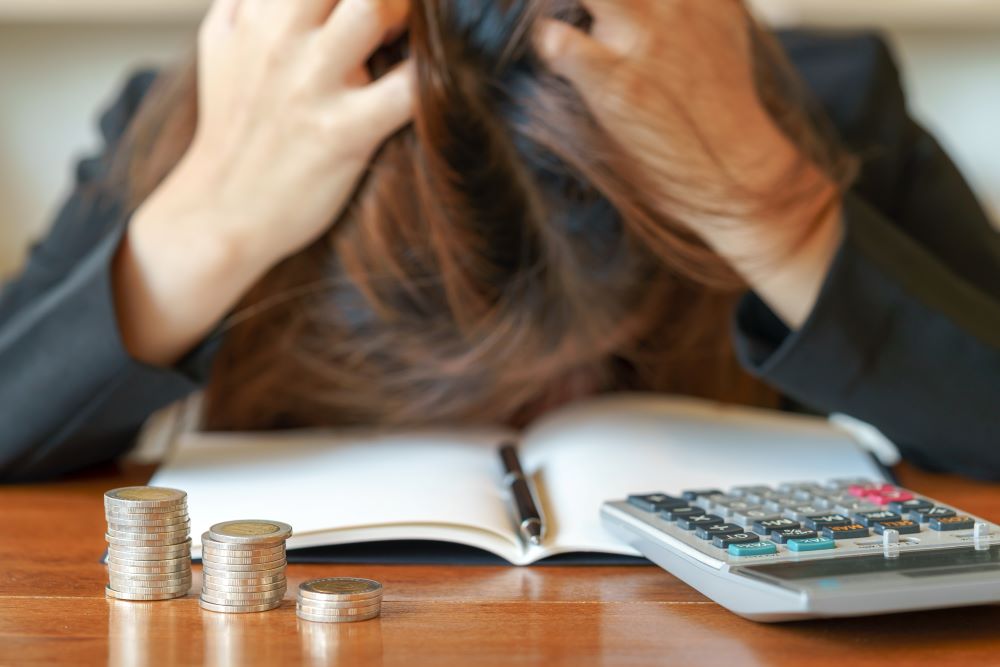 Pessoa com as mãos na cabeça diante de moedas e calculadora, preocupada com finanças e salário família.