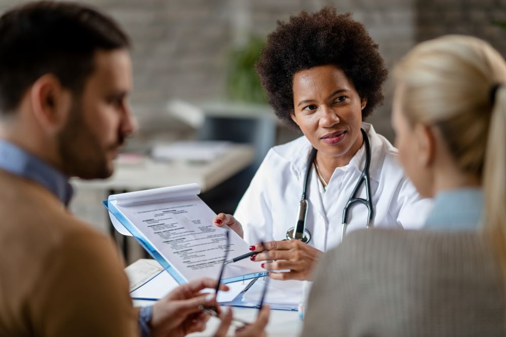 Médica conversa com casal em consulta e apresenta documentos de saúde, representando atendimento médico e benefícios de um plano de saúde empresarial.